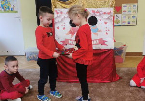 dzieci wręczają sobie kartki walentynkowe, siedzą na dywanie, ubrane w czerwone ubrania 3