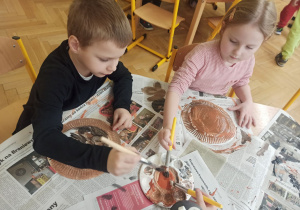 dwoje dzieci maluje farbami telerzyki z papieru siedząc przy stole