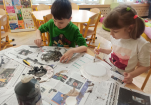chłopiec i dziewczynka malują papierowe talerzyki siedząc przy stole