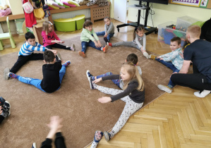 dzieci siedxzą w rozsypoce na dywanie, wykonują ćwiczenie rozciągające