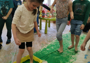 dziewczynka stoi bosymi stopami w pojemniku z zieloną farbą, w tle inne dzieci