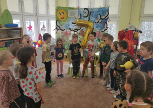 dzieci stoją w kole na dywanie i śpiewają urodzinową piosenkę, na wprost stoją jubilaci, między nimi złoty balon z helem przedstawiający cyfrę 7