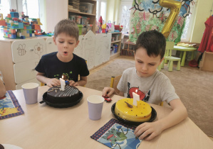 jubilaci siedzą przy stole, przed nimi stoją dwa torty, chłopcy zdmuchują świeczki