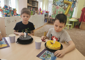jubilaci siedzą przy stole, przed nimi stoją dwa torty, chłopcy zdmuchują świeczki