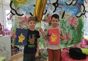 dwaj jubilaci stoją na dywanie, trzymają w ręku złoty balon z helem w kształcie cyfry 7