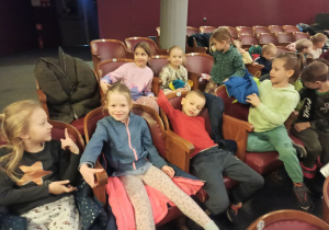 kilkoro dzieci siedzi na krzesłach w teatrze i uśmiechają się