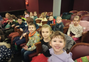 kilkoro dzieci siedzi na krzesłach w teatrze i uśmiechają się