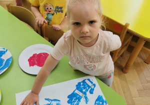 Dziecko odciska kształt dłoni pomalowaną kolorową farbą na białaj kartce papieru.