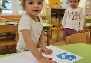 Dziecko odciska kształt dłoni pomalowaną kolorową farbą na białaj kartce papieru.