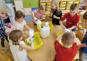 dzieci stoją dookołą stolika i przesypują mąkę ziemniaczaną do żłótych pojemników