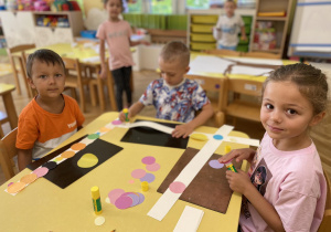 Dzieci siedzą przy stole i przyklejają kolorowe koła do paska