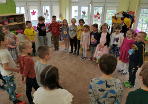 dzieci stoją w kole na dywanie i śpiewają piosenkę urodzinową