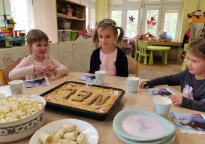 trzy dziewczynki siedzą przy stole i patrzą na ciasto urodzinowe