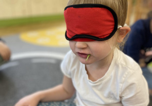 dziecko siedzi na dywanie z opaską na oczach i próbuje rozpoznać warzywo