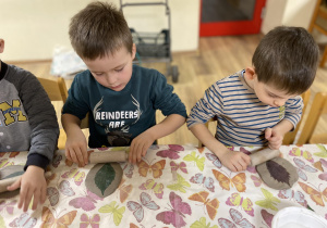 dzieci siedzą przy stole i wałkują glinę wałkiem