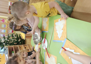 Dzieci smarują choinkę klejem, posypują kaszą manną, malują zieloną farbą oraz dekorują brokatem i cekinami.
