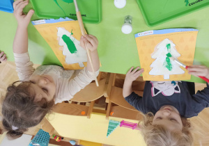 Dzieci smarują choinkę klejem, posypują kaszą manną, malują zieloną farbą oraz dekorują brokatem i cekinami.